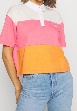 Personne portant un polo color-block blanc, rose et orange avec un jean bleu clair, debout avec une main derrière le dos.
