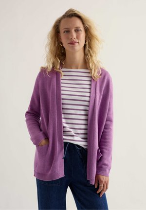 Šviesiaplaukė moteris su auksiniais apvaliais auskarais, purpuriniu megztuku dėvinti baltą ir purpurinę dryžuotą marškinių palaidinę ir tamsiai mėlynas kelnes, stovinti su viena ranka kišenėje.