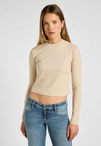Beige ribbet langærmet top med rund halsudskæring, cropped design, diskret logo detalje, parret med blå jeans. Glat tekstur.