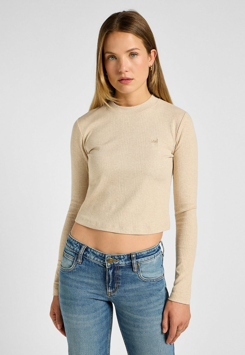 Beige ribbet langærmet top med rund halsudskæring, cropped design, diskret logo detalje, parret med blå jeans. Glat tekstur.