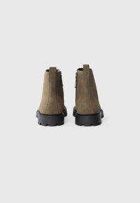 Botas de tobillo de ante verde oliva con una suela de goma negra gruesa, cremallera lateral y detalle de asa para facilitar su uso. Diseño limpio y minimalista.