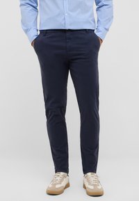 Marineblaue Baumwoll-Chinos mit schlanker Passform, ausgestattet mit Seitentaschen und einer glatten Textur, kombiniert mit weißen und beigen Sneakers.
