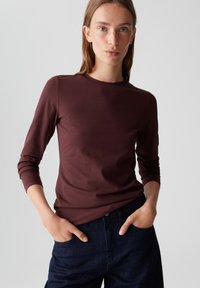 Bordeaux lang shirt met lange mouwen van zachte stof en een ronde hals, gecombineerd met donkere jeans; ontspannen pasvorm met opgerolde mouwen.