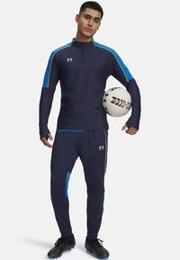 Marineblauw sportensemble met blauwe en gele accenten. Voorzien van een ritssluiting bij de kraag en taps toelopende broekspijpen. Speler houdt een gestructureerde voetbal vast.