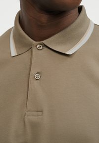 Κοντινό πλάνο ενός ατόμου που φοράει ένα μπεζ πόλο πουκάμισο με ριγέ λευκό και μπεζ γιακά και δύο κουμπιά κλειστά.