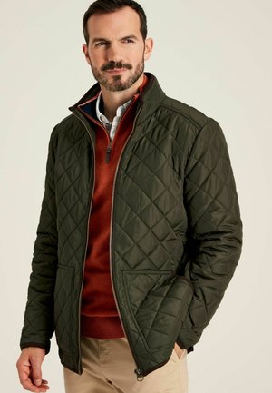 Quilted green jacket with a high collar, featuring a two-way zip, side pockets, and a smooth texture, layered over a rust-colored sweater.
