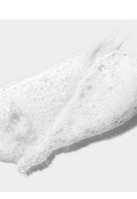 Mousse blanche texturée avec de petites bulles, présentée en forme fluide sur un fond clair, évoquant un produit tel qu'un nettoyant ou un savon.