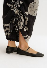 Černé kožené baletky s špičatou špičkou, které mají štíhlý pásik kolem kotníku upevněný ozdobným kovovým kroužkem. Hladký povrch.