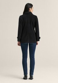 Svart rock med figurnära design, krage och knappklädda manschetter, kombinerad med blå jeans och ankelboots. Slät textur och skräddarsydd silhuett.