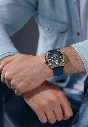 Montre analogique avec un cadran noir, des accents dorés et un boîtier gris massif. Dotée d'un bracelet en caoutchouc bleu-vert et de plusieurs cadrans pour diverses fonctions.
