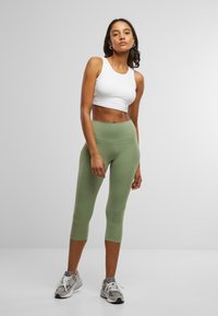 Geknipte witte tanktop en olijfgroene leggings tot halverwege het onderbeen. De leggings hebben een gladde textuur en een hoge tailleband, gecombineerd met grijze sport sneakers.