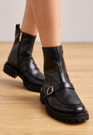 Černé kotníkové boty z hladké kůže, s plochou podrážkou, ozdobou ve formě přezky a zlatým zipem. Masivní vzorek podešve pro lepší přilnavost.