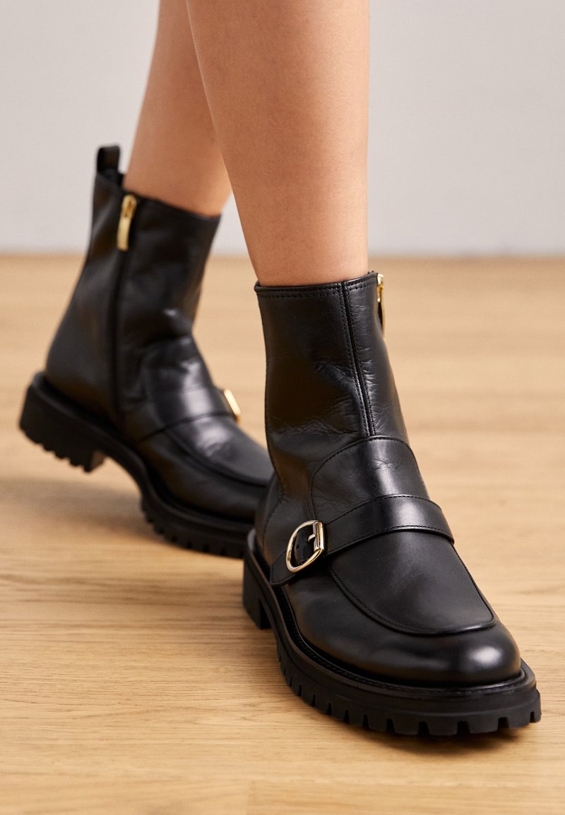 Bottines en cuir noir à texture lisse, dotées d'une semelle plate, d'un détail boucle et d'un accent zip doré. Semelle crantée épaisse pour une meilleure adhérence.