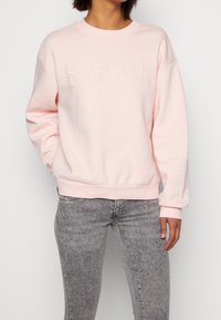 Felpa rosa chiaro con testo in rilievo "SEOUL", collo a giro, spalle scese e polsini e orlo a costine, abbinata a jeans skinny grigi.