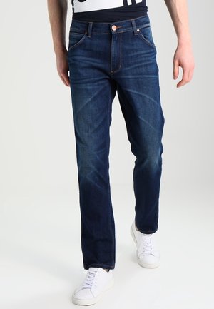 Man met donkerblauwe spijkerbroek en witte sneakers staat voor een effen lichte achtergrond.