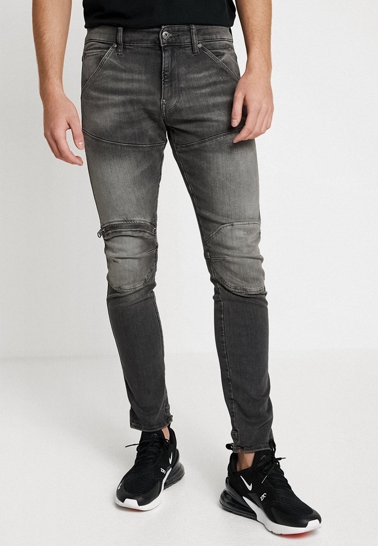 Jeans de corte slim en gris oscuro con paneles texturizados en las rodillas y un ligero desvanecimiento, combinados con zapatillas deportivas negras. Detalles desgastados cerca del dobladillo.