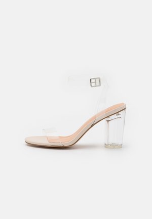 Sandale à talons hauts beige avec une sangle transparente pour les orteils, une sangle de cheville avec une boucle en argent, et un talon épais transparent sur fond blanc.