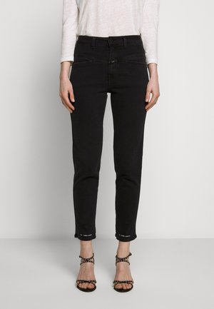 Person trägt schwarze, hoch taillierte Skinny-Jeans und schwarze, mit Nieten verzierte High-Heel-Sandalen und steht vor einem schlichten hellen Hintergrund.