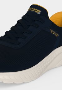 Αθλητικό παπούτσι Skechers slip-in σε ναυτικό μπλε χρώμα με μαύρα κορδόνια, κίτρινη επένδυση, λεπτομέρειες με λογότυπο και παχιά λευκή υφή σόλας.