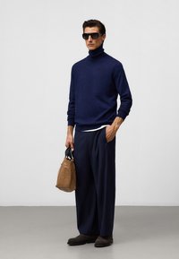 Marinefarbener Rollkragenpullover, locker sitzende dunkle Hose, schwarze Sonnenbrille und braune strukturierte Tasche verstärken den lässigen Look. Dunkle Ankle Boots runden das Outfit ab.