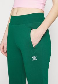 Zelené bavlněné tepláky s bočními kapsami, elastickým pasem a bílým logem Adidas na dolní části levé nohy. Hladká textura.