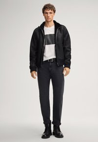 Zwarte bomberjack met een zachte kraag, wit T-shirt met zwart grafisch ontwerp, donkere jeans en zwarte veterschoenen.