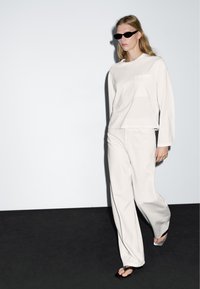 Hellgraues Langarmshirt mit Tasche und passenden weiten Hosen, kombiniert mit schwarzen Sonnenbrillen und Flip-Flop-Sandalen. Minimalistisches Design.