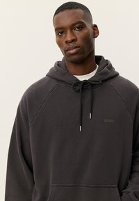 Σκούρα γκρι φόρμα με κουκούλα με τσέπη καγκουρό, κορδόνι κουκούλας και μανίκια ραγκλάν. Διαθέτει μικρό, καφέ λογότυπο της μάρκας. Μαλακή υφή υφάσματος.