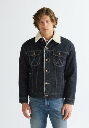Man with curly hair stands facing forward, wearing a dark denim sherpa jacket with a white fleece collar and blue jeans against a plain background.