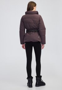 Mujer vestida con una chaqueta acolchada de color púrpura oscuro y cintura ajustada, leggings negros y botas forradas de piel negra, de pie contra un fondo liso.