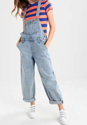 Personne portant un t-shirt rayé bleu et orange avec une salopette ample en jean bleu clair et des baskets blanches, les mains dans les poches, debout sur un sol blanc.