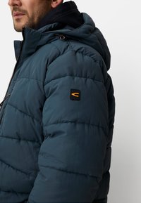 Tyrkisblå dunjakke med lynlås foran, hætte, tekstureret quiltet mønster og en orange logo-patch på venstre ærme.