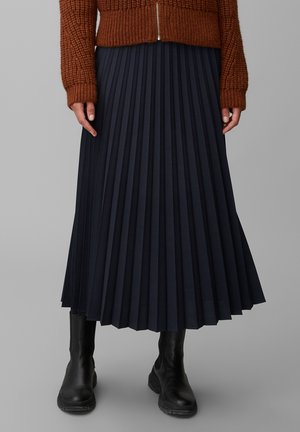 Femme portant un pull en tricot marron, une jupe midi plissée bleu marine et des bottes en cuir noir, debout devant un fond gris.