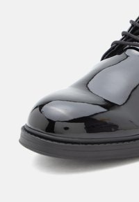 Pantof din piele în culorile negru lac, cu finisaj lucios, vârf rotund și talpă căptușită de culoare neagră. Are șireturi texturate și un design neted în ansamblu.