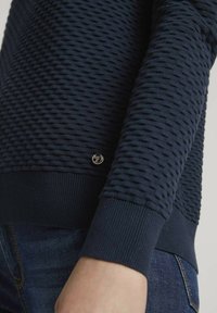 Sweatshirt tricoté bleu marine avec un motif texturé, des poignets côtelés et un petit logo métallique près de l'ourlet, associé à un jean sombre.