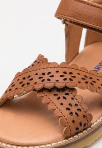 Brune lær sandaler med utskårne blomsterdesign og bølgete kanter. Sålen er lett og teksturert, noe som fremhever komfort og stil.