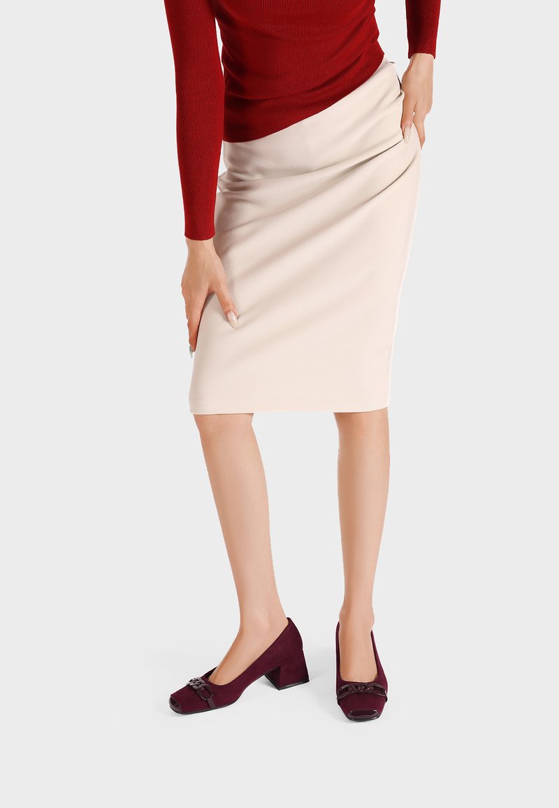 Gonna a matita beige abbinata a scarpe basse bordeaux con un dettaglio a fiocco. Maglietta rossa a maniche lunghe texturizzata parzialmente visibile.