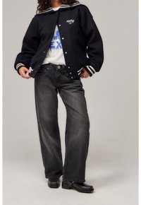 Black varsity jacket with white stripes, graphic white t-shirt, and wide-leg black jeans. Outfitted with black ankle boots.