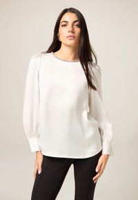 Hellbeige Bluse mit langen Ärmeln, plissierten Schultern und einer strukturierten Halslinie. Der Stoff wirkt glatt und fällt locker über schwarze Hosen.