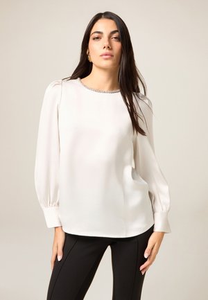 Light beige blouse with long sleeves, pleated shoulders, and a textured neckline. Fabric appears smooth and drapes loosely over black pants.
