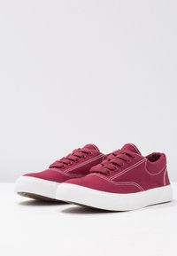 Scarpe da ginnastica in tela color granata con suole e lacci bianchi in gomma. Cuciture bianche evidenti e design con punta arrotondata.