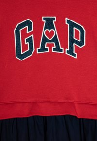 Sweatshirt roșu cu un logo "GAP" albastru marin, având o formă de inimă în locul literei "A". La bază, are o fustă plisată albastră marin.