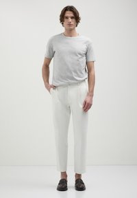 Uomo con capelli di lunghezza media che indossa una t-shirt grigia chiara, pantaloni bianchi su misura e mocassini marroni scuri, in piedi contro uno sfondo neutro.