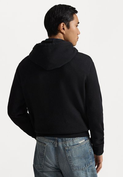 Vyras, apsirengęs juoda kapo tipo bliuzonu ir mėlynomis džinsomis, šonu atsisukęs su dalimi nugaros link kameros, prieš paprastą baltą foną.