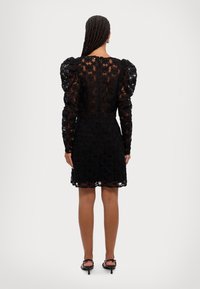 Svart spetsklänning med blommönster, långa ärmar och puffiga axlar. Knytelekslängd, genomskinlig med solid underdel, kompletterad med remsandaler.