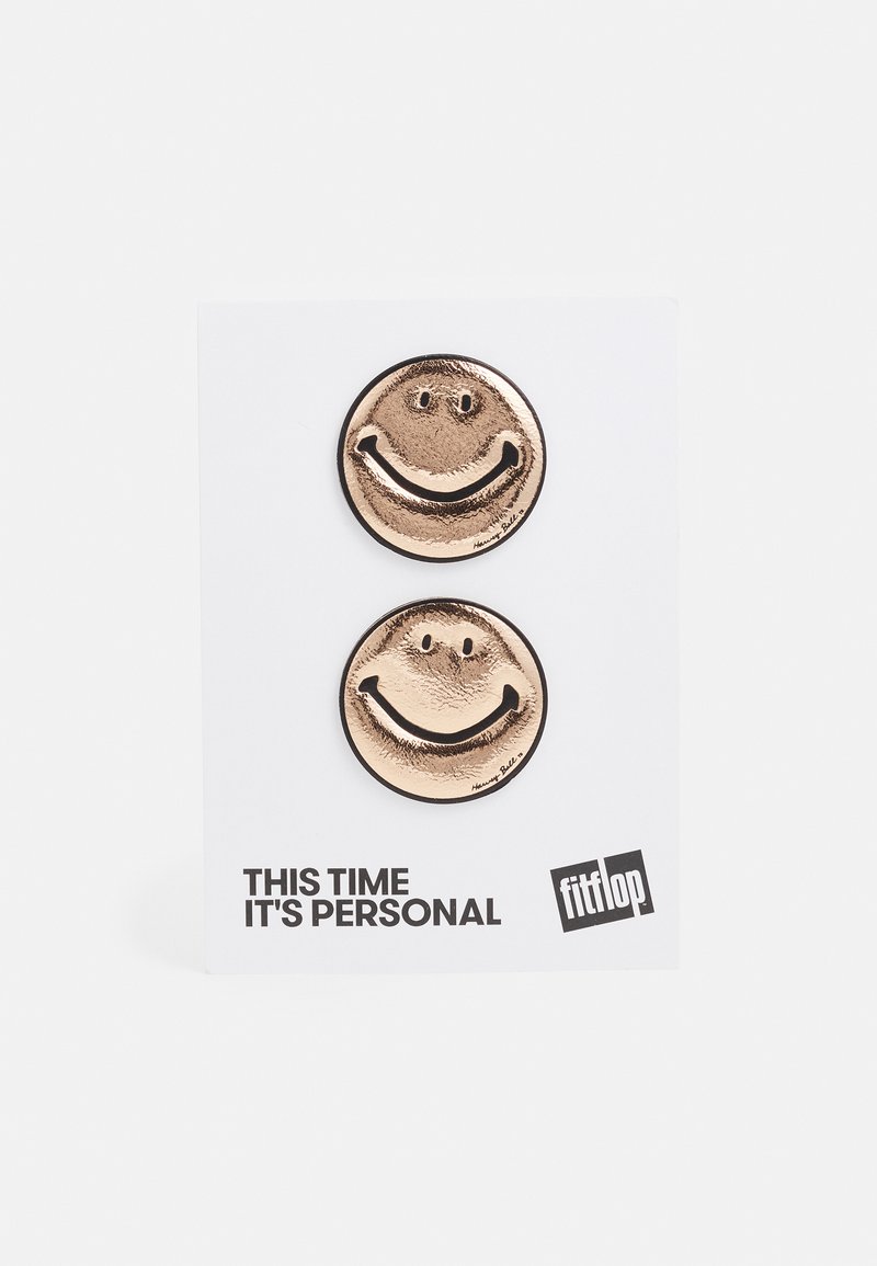 Botões de rosto sorridente em metal dourado sobre um cartão branco, apresentando uma superfície texturizada e detalhes pretos. O texto diz "Desta vez é pessoal."