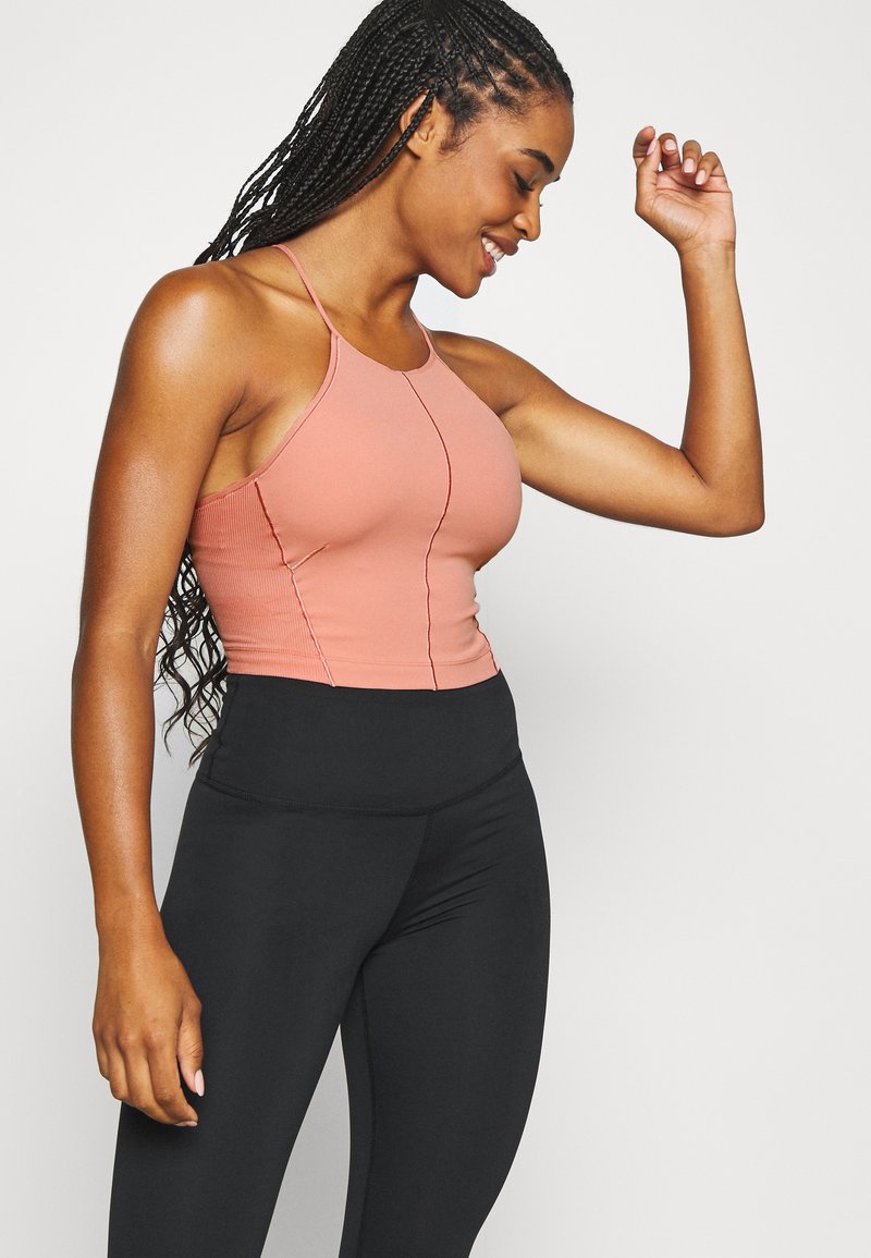 Camiseta de tirantes finos de color melocotón con canalé; diseño ajustado con detalles de costura en contraste. Combinada con leggings negros de cintura alta.