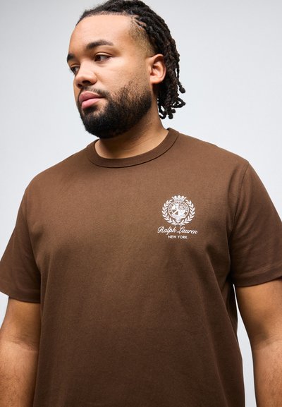 Άνδρας με γένια και κοτσίδες dreadlocks που φοράει καφέ μπλουζάκι Ralph Lauren New York, κοιτάζοντας προς τα αριστερά, μπροστά σε απλό φόντο.