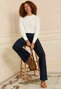 Maglia bianca a maniche lunghe con scollo in pizzo, jeans svasati scuri, sandali beige con cinturino e una piccola borsa a strappo marrone appoggiata sulle ginocchia.