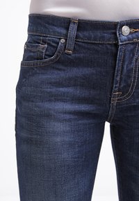 Jeans de mezclilla oscura con una textura suave, que presentan un diseño clásico de cinco bolsillos, costuras en contraste y un cierre de botón de metal en la cintura.